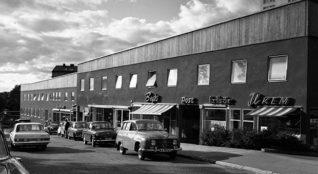Svartvit bild på gata med parkerade bilar och butiksfasader 1965.